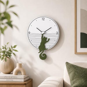 Op de afbeelding: Een witte wandklok met zwarte wijzers en een groen katten silhouet dat aan de wijzerplaat hangt. De klok heeft een minimalistisch ontwerp met zwarte lijnen en cijfers op de posities 12, 3, 6 en 9 uur. De staart van de kat vormt een decoratieve krul.