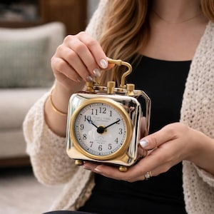Könnte beinhalten: Eine quadratische, silberfarbene Uhr mit goldfarbenem Zifferblatt und Akzenten. Das Zifferblatt zeigt schwarze Ziffern und Zeiger. Die Uhr hat einen Griff und zwei Knöpfe oben. Die Worte "Daniel Wellington Clock Company" sind auf dem Zifferblatt aufgedruckt.