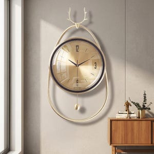 May include: A gold-toned wall clock with a circular face and a black border. The clock features a pendulum and is suspended within a gold frame with a deer antler design at the top. The clock hands point to 10:15.
