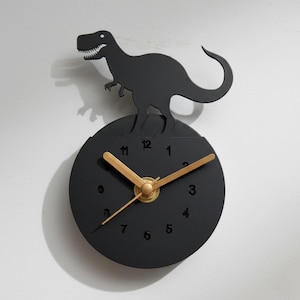 May include: A black wall clock featuring a dinosaur silhouette above the clock face. The clock has a round black face with gold-colored hands and numbers. The dinosaur is a Tyrannosaurus Rex.