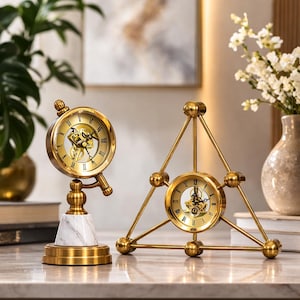 May include: Two decorative gold-colored clocks. One clock is round, mounted on a white marble base. The other clock is triangular, with a clock face in the center. Both clocks have Roman numerals and visible clock mechanisms.