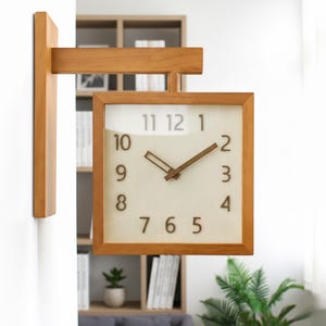 May include: A double-sided wooden wall clock with a square face. The clock has a light beige background with brown numerals and hands. The clock is mounted on a wooden frame and arm, and is attached to a wall.