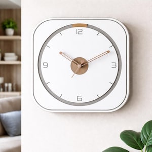 Op de afbeelding: Vierkante witte wandklok met een minimalistisch ontwerp. De klok heeft een witte wijzerplaat met zwarte cijfers op 3, 6, 9 en 12 uur, en goudkleurige wijzers. De klok heeft een witte lijst en de algehele esthetiek is modern en strak.