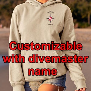 May include: A beige hooded sweatshirt with a front pocket and the text "Customizable with divemaster name" in red. The sweatshirt has a small emblem with the word "Divemaster" and a name below it. The person is wearing denim shorts.