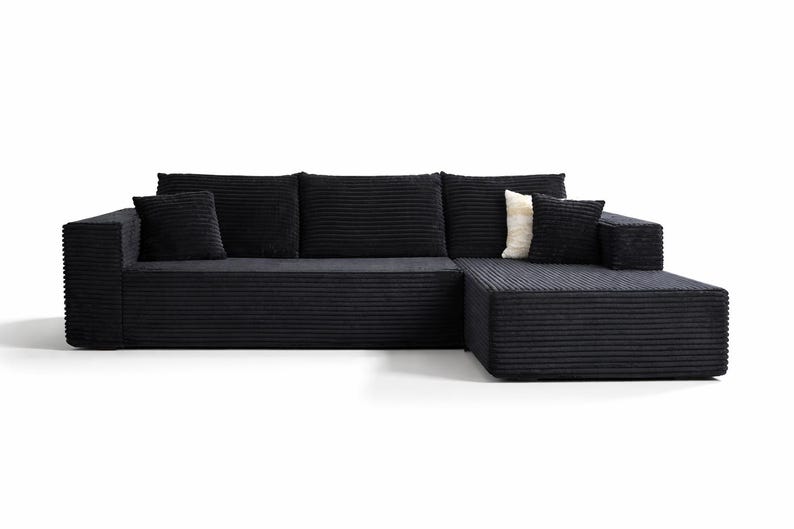 K&ouml;nnte beinhalten: Ein modernes schwarzes Ecksofa mit Cordstruktur. Das Sofa hat eine Chaiselongue auf der rechten Seite und enth&auml;lt drei gro&szlig;e R&uuml;ckenkissen und zwei kleinere quadratische Kissen. Ein cremefarbenes Kissen sorgt f&uuml;r Kontrast.