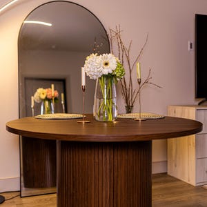 May include: A round, dark brown wooden dining table with a fluted base. The table is set with a vase of white and green flowers, two candles, and decorative placemats. A large arched mirror reflects the table and decor.
