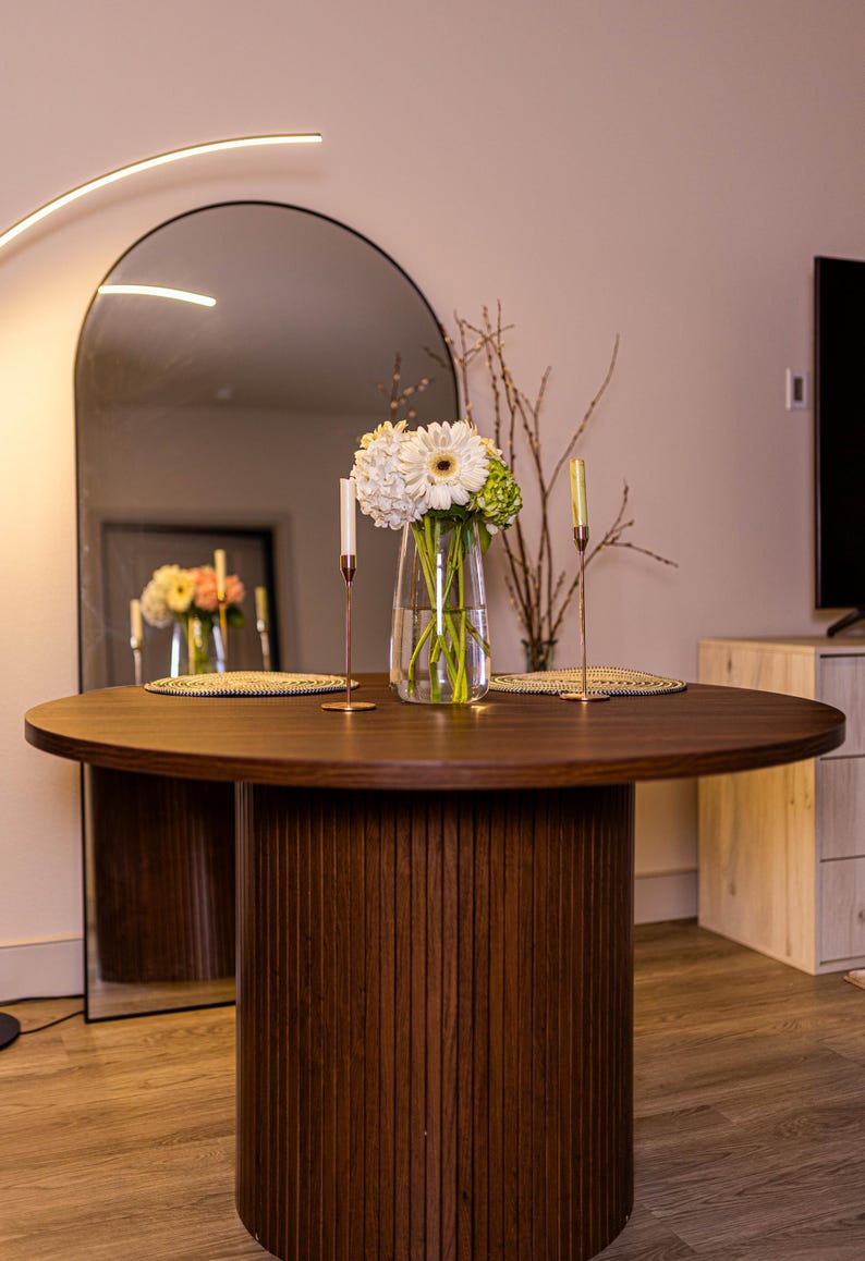 May include: A round, dark brown wooden dining table with a fluted base. The table is set with a vase of white and green flowers, two candles, and decorative placemats. A large arched mirror reflects the table and decor.