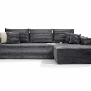 K&ouml;nnte beinhalten: Dunkelgraues Ecksofa aus Cordstoff. Das Sofa wird von mehreren passenden Kissen und einem flauschigen Kissen begleitet. Ein Buch und ein Glas sind ebenfalls sichtbar.