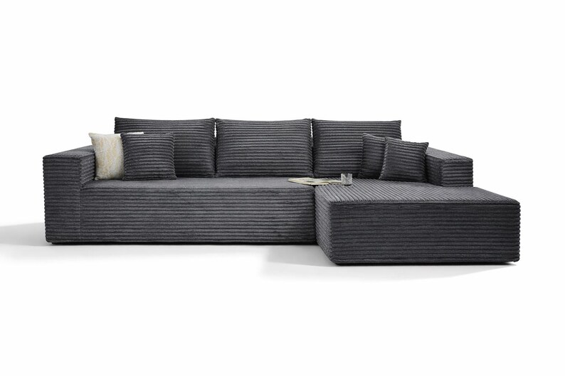 K&ouml;nnte beinhalten: Dunkelgraues Ecksofa aus Cordstoff. Das Sofa wird von mehreren passenden Kissen und einem flauschigen Kissen begleitet. Ein Buch und ein Glas sind ebenfalls sichtbar.
