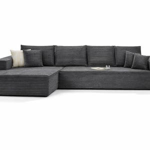 K&ouml;nnte beinhalten: Ein gro&szlig;es, modernes Ecksofa aus dunkelgrauem Cordstoff. Das Sofa hat eine Chaiselongue auf der linken Seite und mehrere passende Kissen. Ein kleines Glas und ein Buch liegen auf dem Sofa. Das Sofa steht vor wei&szlig;em Hintergrund.