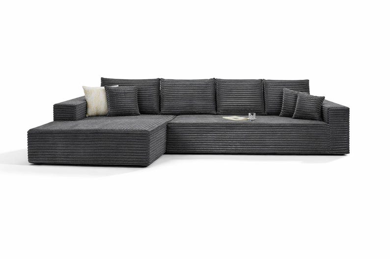 K&ouml;nnte beinhalten: Ein gro&szlig;es, modernes Ecksofa aus dunkelgrauem Cordstoff. Das Sofa hat eine Chaiselongue auf der linken Seite und mehrere passende Kissen. Ein kleines Glas und ein Buch liegen auf dem Sofa. Das Sofa steht vor wei&szlig;em Hintergrund.