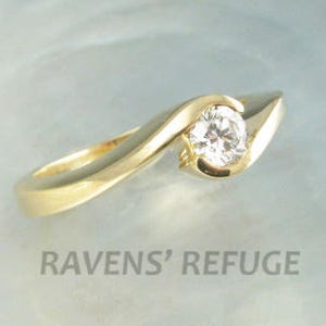 Op de afbeelding: Een gouden ring met een enkele ronde diamant in een gebogen zetting. De ring ligt op een lichtblauwe achtergrond.