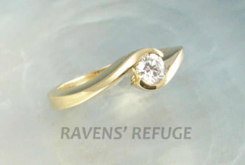 Op de afbeelding: Een gouden ring met een enkele ronde diamant in een gebogen zetting. De ring ligt op een lichtblauwe achtergrond.