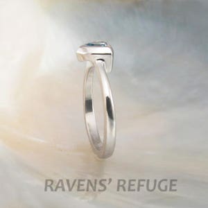May include: A silver ring with a blue gemstone set in a prong setting. The ring is shown from the side.