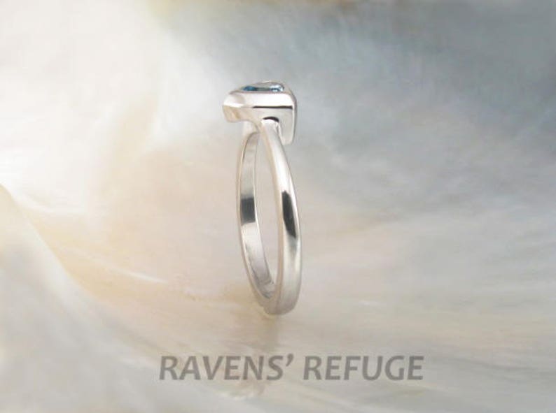 May include: A silver ring with a blue gemstone set in a prong setting. The ring is shown from the side.