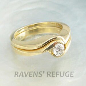 Op de afbeelding: Een paar gouden trouwringen met een enkele ronde diamant die in het midden van de bovenste ring is gezet. De ringen zijn met elkaar verweven en de diamant is gezet in een bezel-setting.