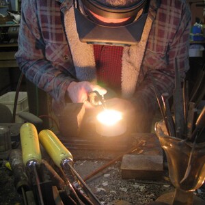 May include: A person wearing a plaid shirt and a welding mask is using a torch to melt metal in a crucible. The metal is glowing orange hot. There are several tools in a vase on the table.