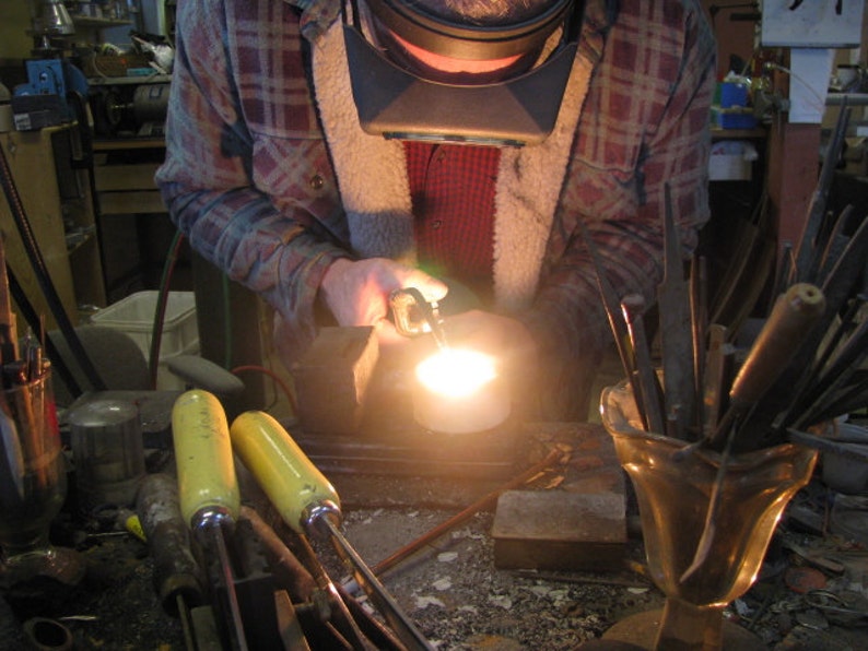 May include: A person wearing a plaid shirt and a welding mask is using a torch to melt metal in a crucible. The metal is glowing orange hot. There are several tools in a vase on the table.