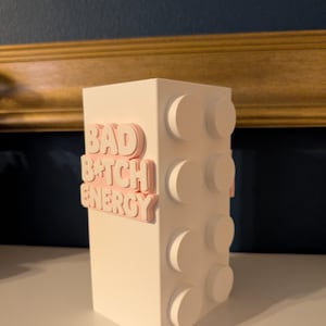 May include: A white, rectangular block-shaped decorative item with raised circular details on one side. The words "BAD B*TCH ENERGY" are in pink, raised lettering on the front. The item is on a white surface.