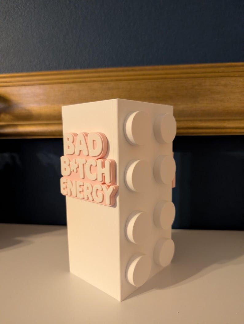 May include: A white, rectangular block-shaped decorative item with raised circular details on one side. The words "BAD B*TCH ENERGY" are in pink, raised lettering on the front. The item is on a white surface.
