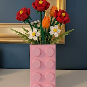 May include: A pink Lego-style vase holds a bouquet of red roses, orange tulips, and white daisies. The flowers have green stems and leaves. The vase is rectangular with eight pink circular studs. A gold-framed picture hangs in the background.