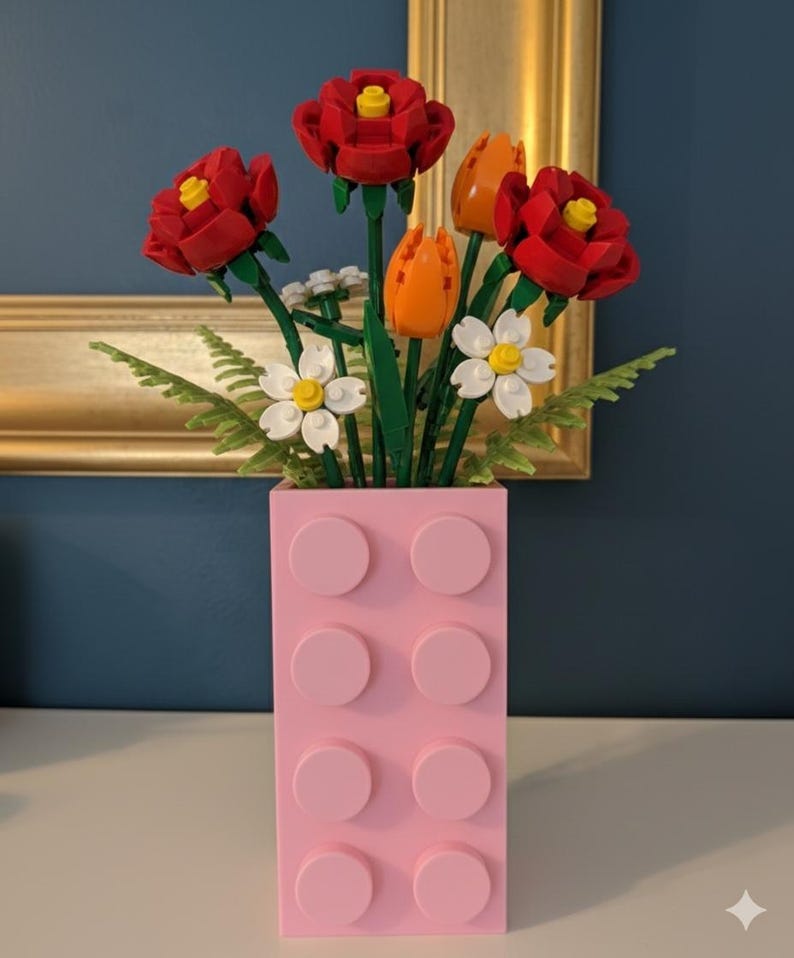 May include: A pink Lego-style vase holds a bouquet of red roses, orange tulips, and white daisies. The flowers have green stems and leaves. The vase is rectangular with eight pink circular studs. A gold-framed picture hangs in the background.