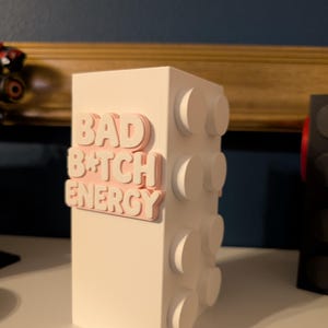 May include: A white rectangular block with raised circular details on the side. The words "BAD B*TCH ENERGY" are in pink, raised lettering on the front of the block. The block is sitting on a white surface.