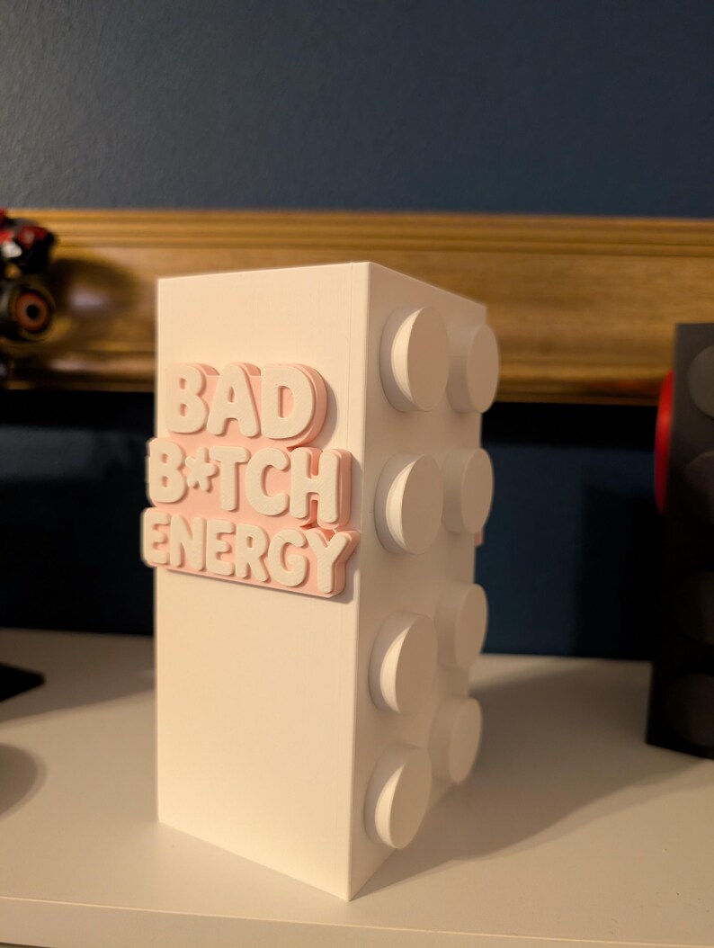 May include: A white rectangular block with raised circular details on the side. The words "BAD B*TCH ENERGY" are in pink, raised lettering on the front of the block. The block is sitting on a white surface.
