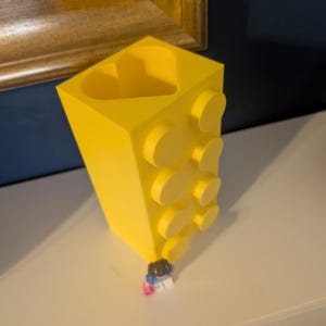 May include: A bright yellow, block-shaped vase with a heart-shaped opening. The vase has several circular studs on the side, resembling a building block. A small Lego figure is at the base of the vase.