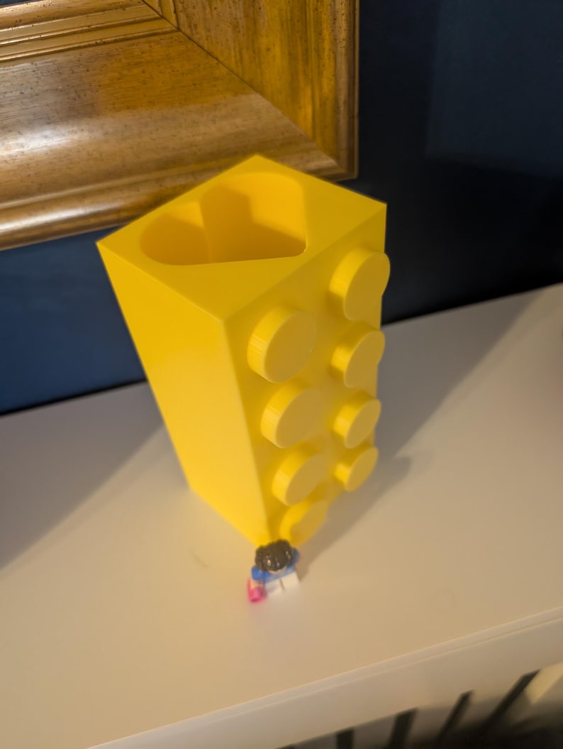May include: A bright yellow, block-shaped vase with a heart-shaped opening. The vase has several circular studs on the side, resembling a building block. A small Lego figure is at the base of the vase.