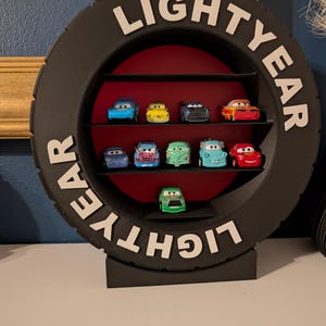 May include: A black tire-shaped shelf unit with the word "LIGHTYEAR" in white lettering. The shelf displays several toy cars in various colors, including blue, yellow, and red. The interior of the shelf is red.