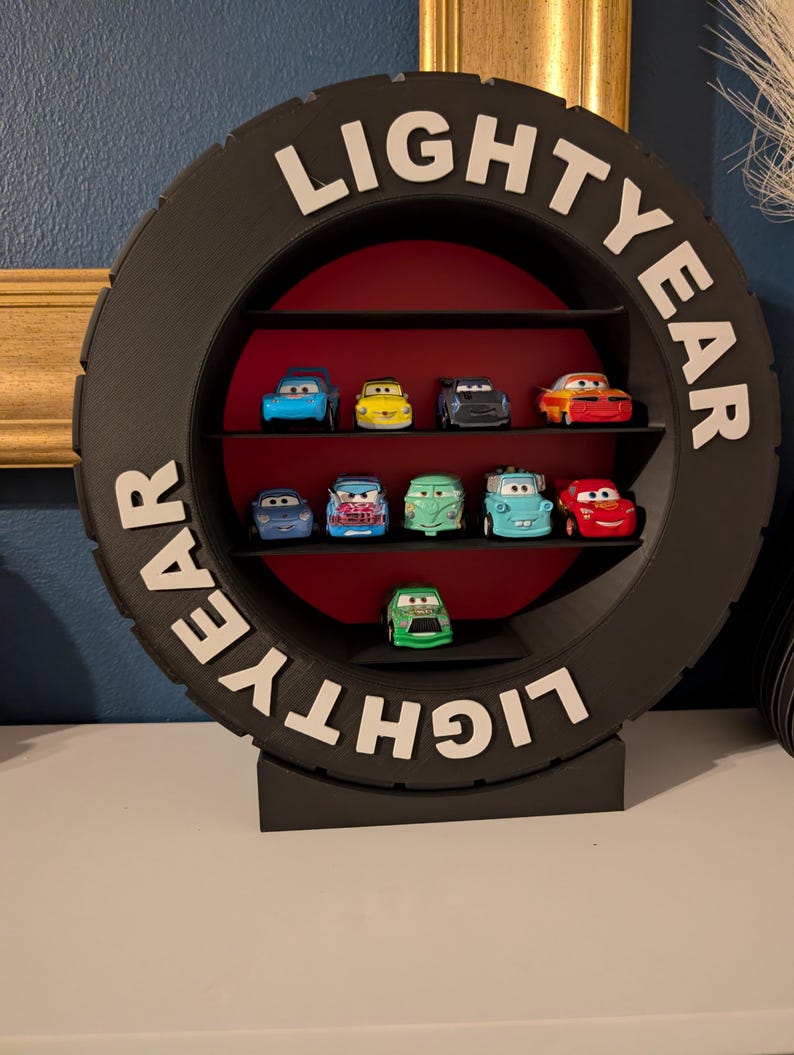 May include: A black tire-shaped shelf unit with the word "LIGHTYEAR" in white lettering. The shelf displays several toy cars in various colors, including blue, yellow, and red. The interior of the shelf is red.
