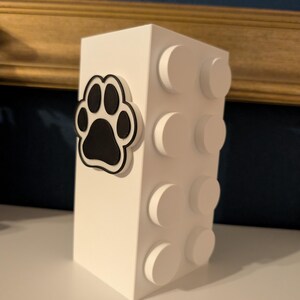 May include: A white, rectangular decorative box with a black paw print design on the front. The side of the box features white, circular, raised details. The box is likely a pet-themed item.