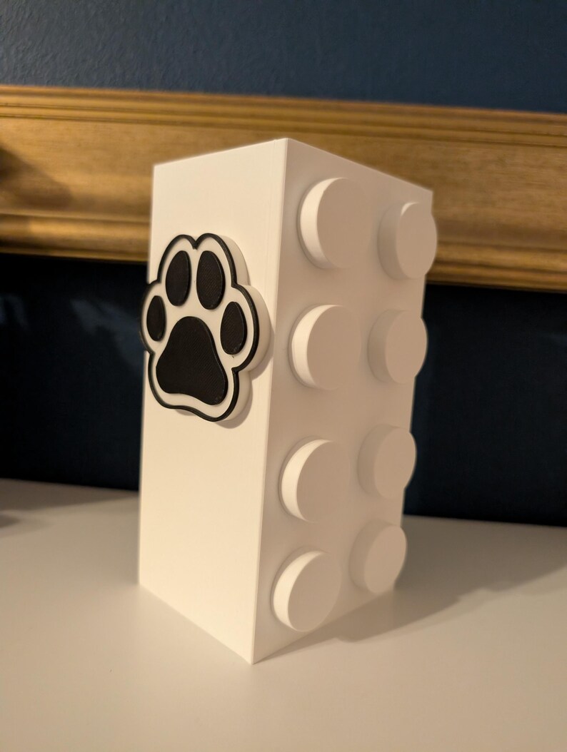 May include: A white, rectangular decorative box with a black paw print design on the front. The side of the box features white, circular, raised details. The box is likely a pet-themed item.