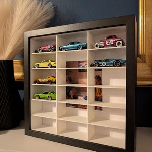 May include: A black framed display case with multiple compartments, showcasing a collection of toy cars. The cars are various colors, including red, blue, yellow, and green. The case is on a white surface, with a dark blue wall in the background.