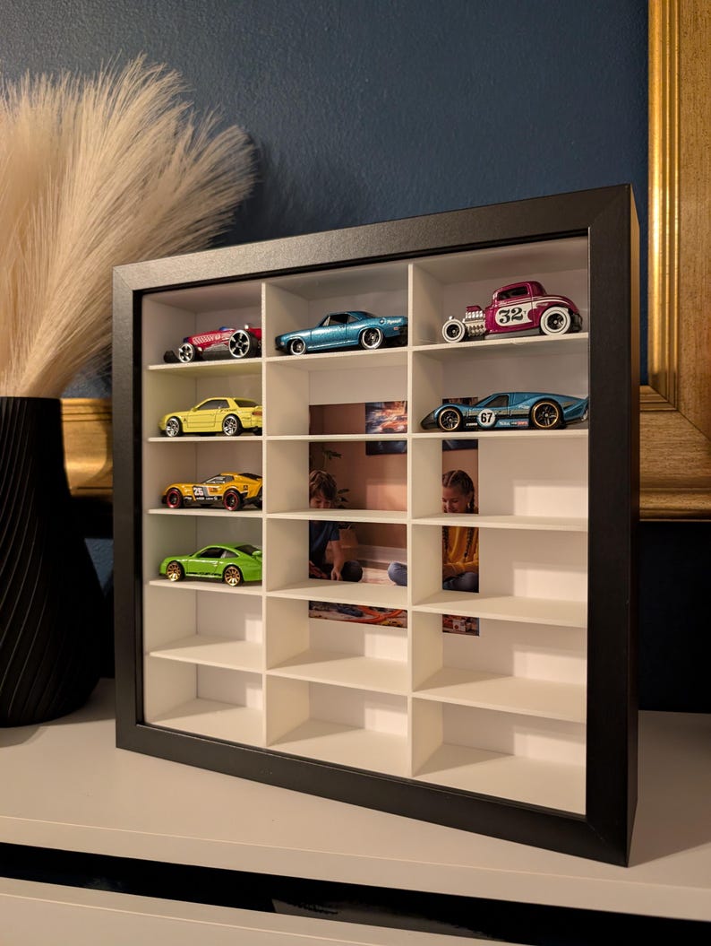 May include: A black framed display case with multiple compartments, showcasing a collection of toy cars. The cars are various colors, including red, blue, yellow, and green. The case is on a white surface, with a dark blue wall in the background.