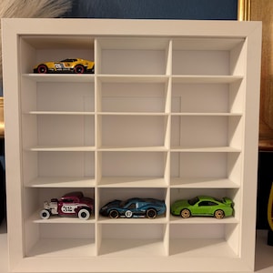 May include: A white, square display case with multiple compartments, showcasing toy cars. The case holds a yellow race car, a maroon vintage car, a blue sports car, and a green sports car. The cars are in various colors and designs, adding a vibrant touch to the display.