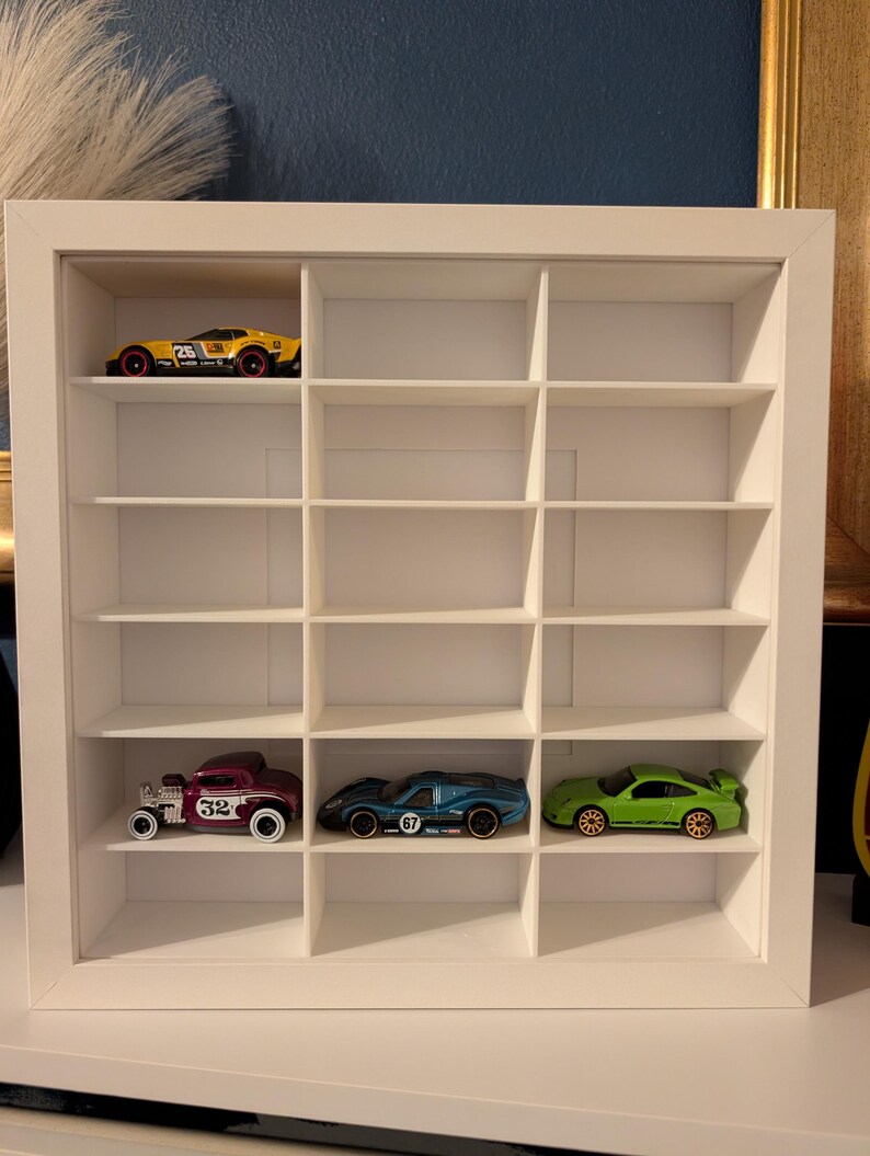 May include: A white, square display case with multiple compartments, showcasing toy cars. The case holds a yellow race car, a maroon vintage car, a blue sports car, and a green sports car. The cars are in various colors and designs, adding a vibrant touch to the display.