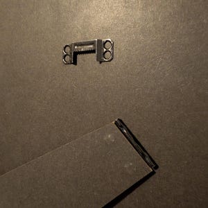 May include: Close-up of the back of a picture frame. A metal sawtooth hanger is attached to the dark gray backing. A black easel back is angled at the bottom, providing support for the frame.