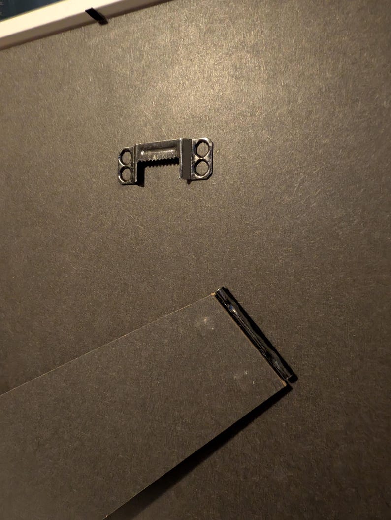 May include: Close-up of the back of a picture frame. A metal sawtooth hanger is attached to the dark gray backing. A black easel back is angled at the bottom, providing support for the frame.