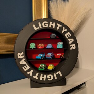 May include: A black, tire-shaped display shelf with the word "LIGHTYEAR" in white lettering. The shelf contains several toy cars of various colors, arranged on red shelves. The display sits on a white surface.