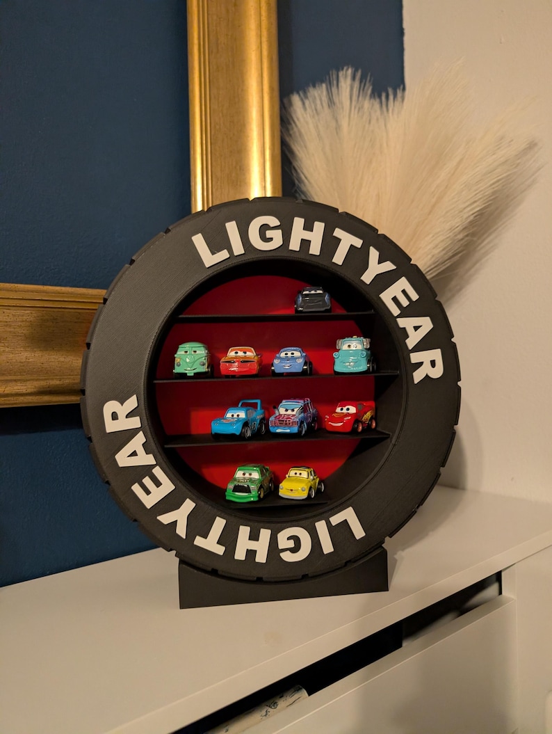 May include: A black, tire-shaped display shelf with the word "LIGHTYEAR" in white lettering. The shelf contains several toy cars of various colors, arranged on red shelves. The display sits on a white surface.