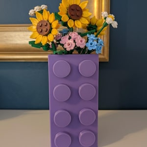 May include: A purple brick-shaped vase filled with colorful Lego flowers, including yellow sunflowers, pink and blue blossoms, and white flowers. The vase has eight circular studs on the front.