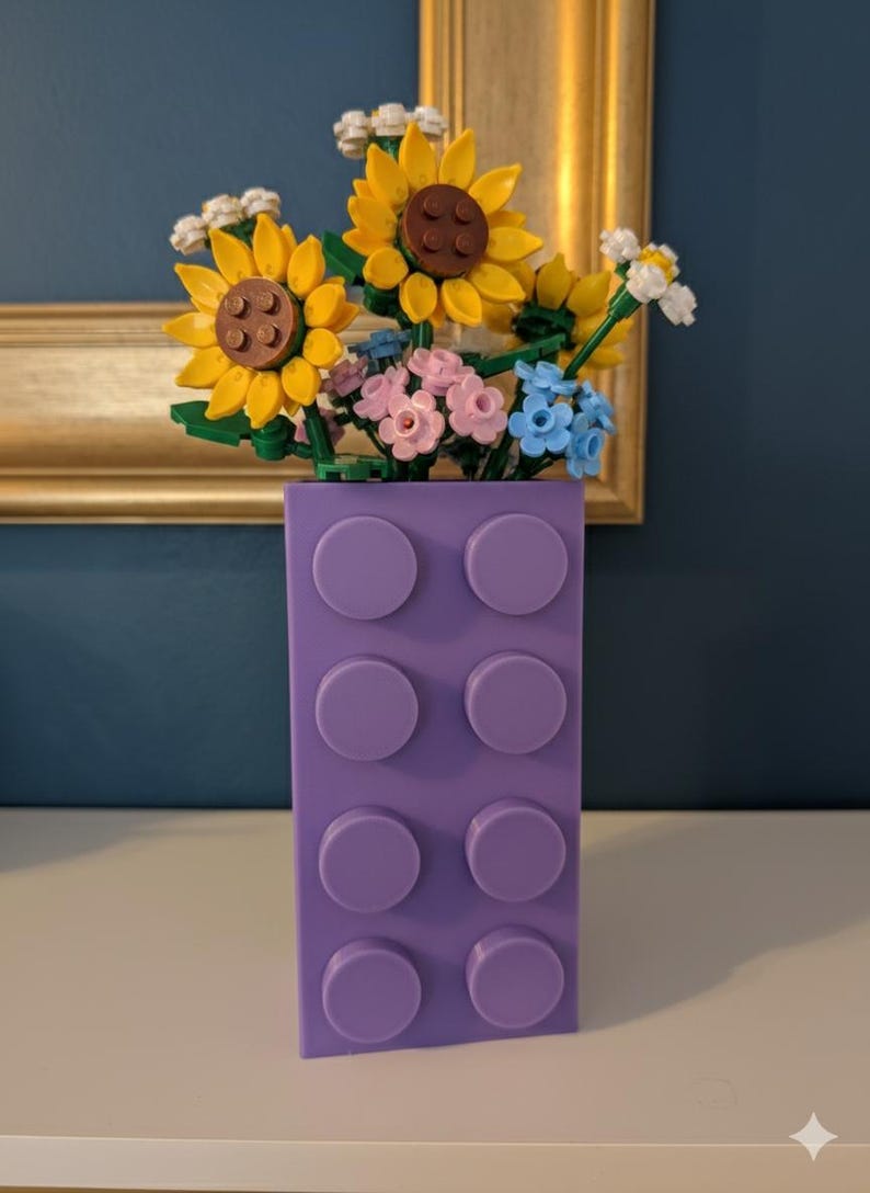 May include: A purple brick-shaped vase filled with colorful Lego flowers, including yellow sunflowers, pink and blue blossoms, and white flowers. The vase has eight circular studs on the front.
