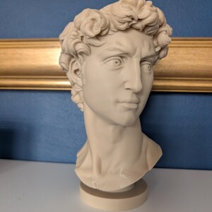 May include: A cream-colored bust of a man with curly hair, a stern expression, and a detailed face. The sculpture is on a round base and set against a blue wall with a gold-colored frame.