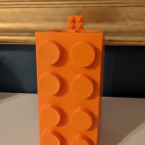 Op de afbeelding: Fel oranje container in de vorm van een bouwsteen, met acht ronde noppen aan de voorkant en een kleiner blokje bovenop. De container heeft een fel oranje kleur en staat op een wit oppervlak.