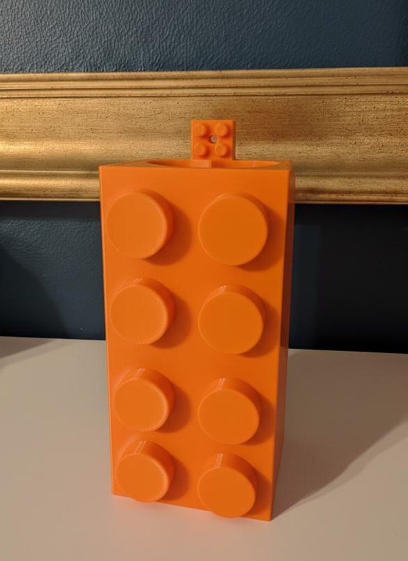 Op de afbeelding: Fel oranje container in de vorm van een bouwsteen, met acht ronde noppen aan de voorkant en een kleiner blokje bovenop. De container heeft een fel oranje kleur en staat op een wit oppervlak.