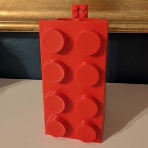 Op de afbeelding: Een felrode, rechthoekige blokvormige container met acht verhoogde cirkelvormige noppen aan de voorkant. Een kleiner blok met vier noppen is aan de bovenkant bevestigd. De container is waarschijnlijk een pennenhouder of bureau-organizer.