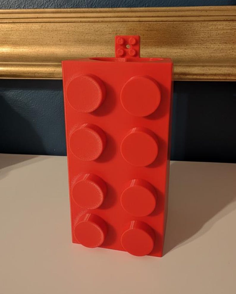 Op de afbeelding: Een felrode, rechthoekige blokvormige container met acht verhoogde cirkelvormige noppen aan de voorkant. Een kleiner blok met vier noppen is aan de bovenkant bevestigd. De container is waarschijnlijk een pennenhouder of bureau-organizer.