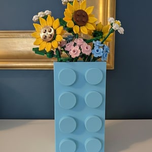 May include: A light blue vase made of interlocking blocks holds a bouquet of colorful flowers. The flowers include yellow sunflowers, pink and blue blossoms, and white accents. The vase is on a white surface.